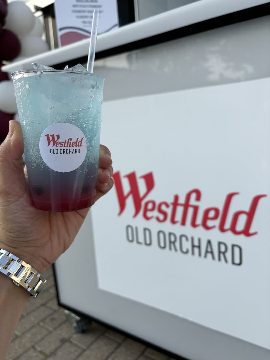 A layered blue Abu Sauce mocktail held in front of the Westfield Old Orchard sign