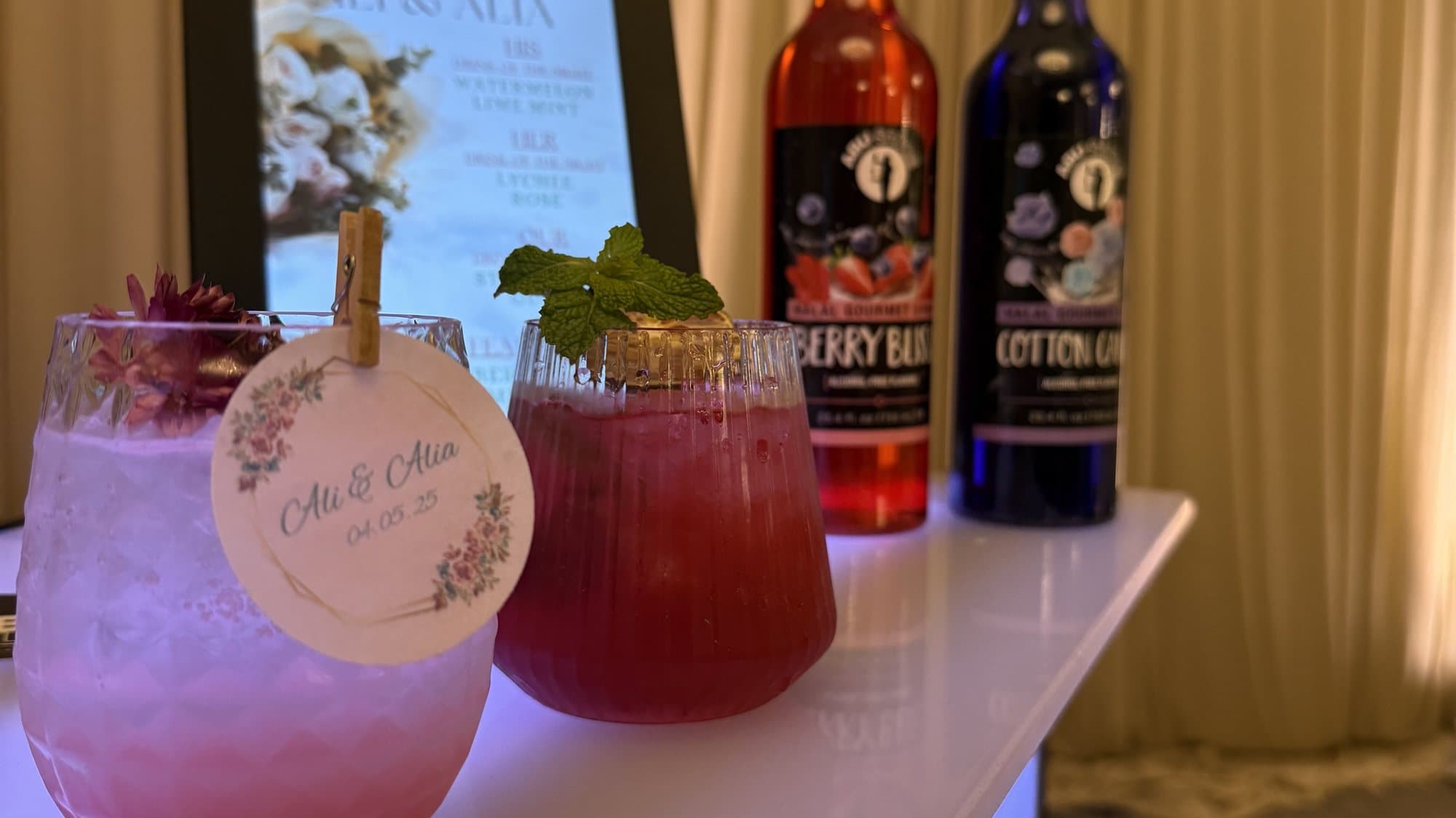 Two garnished mocktails with custom 'Ali & Alia' wedding drink tags beside Abu Sauce syrup bottles on a lit bar