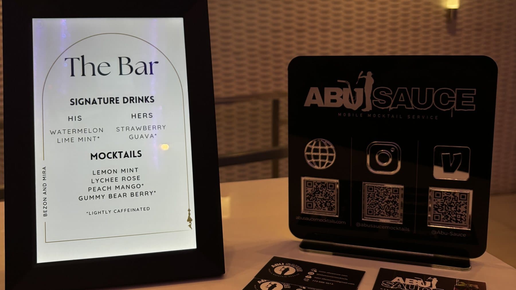 A framed 'The Bar' signature drinks menu beside Abu Sauce branded cards and a QR stand