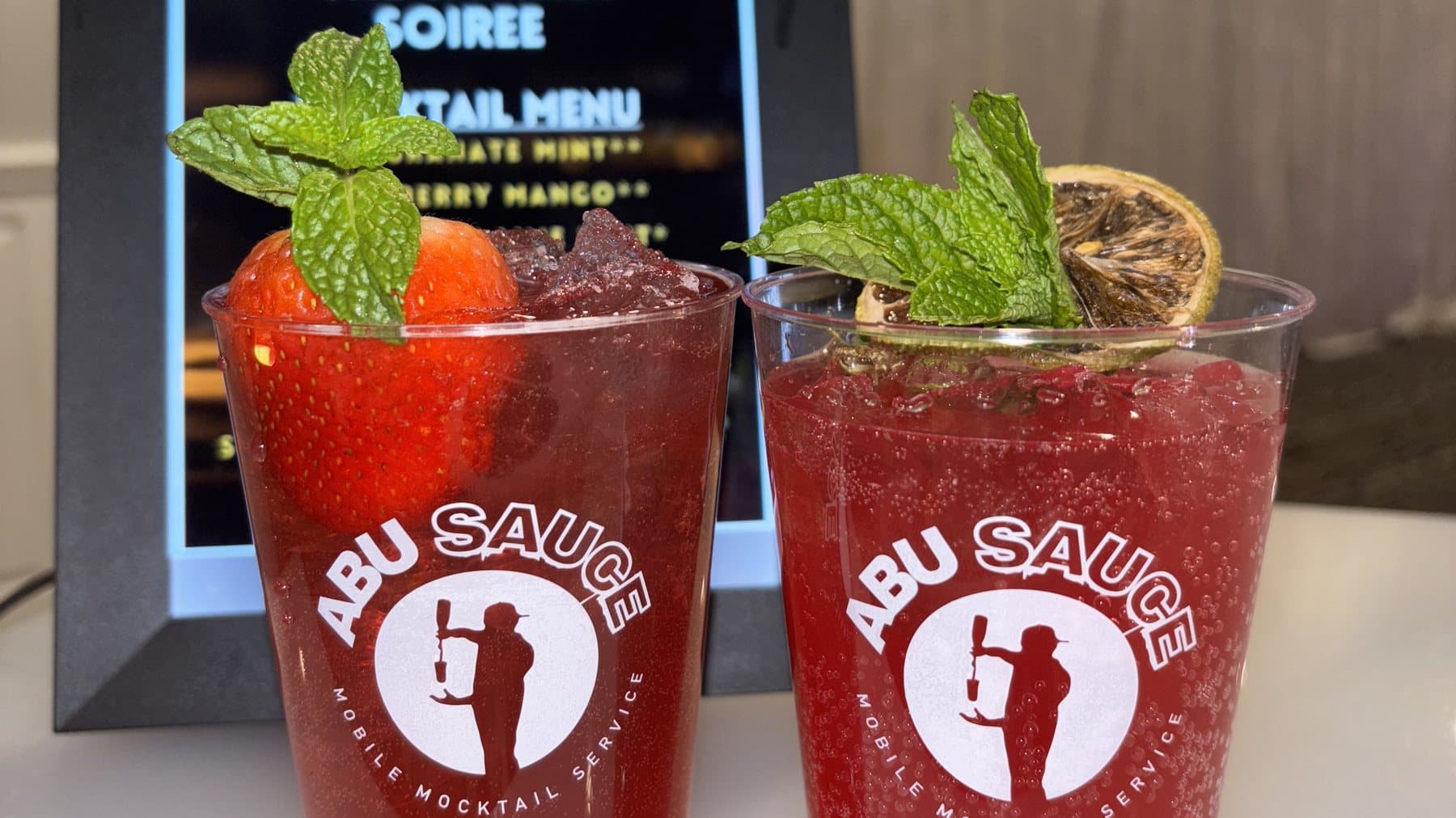 Two red Abu Sauce mocktails garnished with mint and dried orange in front of a Sahara Soirée mocktail menu
