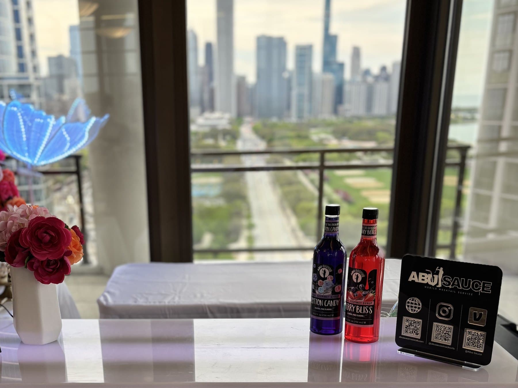 A lit Abu Sauce mocktail bar with syrup bottles and flowers in front of floor-to-ceiling windows over the Chicago skyline